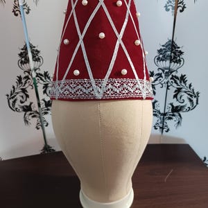 May include: A burgundy-colored hat with a diamond pattern created by silver ribbon. The hat is adorned with small, round, white pearls at the intersections of the ribbon. The base of the hat features a silver lace trim. The hat is displayed on a mannequin head.