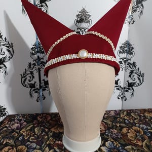 May include: A burgundy jester hat with two pointed tips. The hat is trimmed with white and gold decorative trim and a central pearl-like button. The hat is displayed on a mannequin head against a floral patterned background.