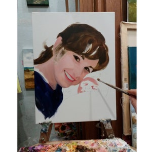 May include: A portrait painting on a white canvas, depicting a smiling person with brown hair and a blue top. The painting is on an easel, with a palette of colors in the foreground. A paintbrush is held near the canvas.