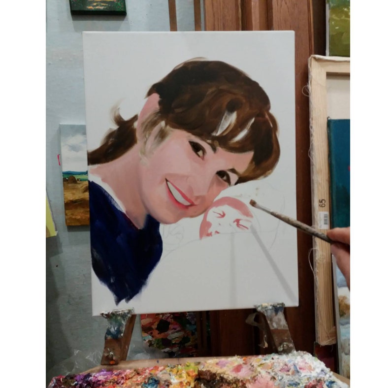 May include: A portrait painting on a white canvas, depicting a smiling person with brown hair and a blue top. The painting is on an easel, with a palette of colors in the foreground. A paintbrush is held near the canvas.