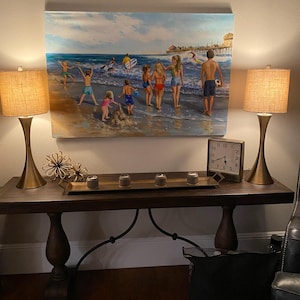 May include: A beach scene painting hangs above a dark wood console table. Two gold-toned lamps with tan shades flank the painting. The table holds a tray with candles and a clock. A black bag sits below.