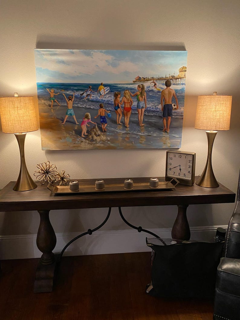 May include: A beach scene painting hangs above a dark wood console table. Two gold-toned lamps with tan shades flank the painting. The table holds a tray with candles and a clock. A black bag sits below.