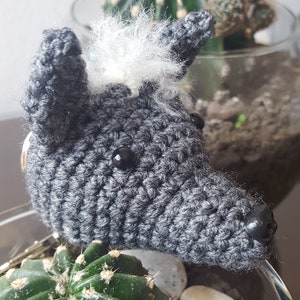 May include: A gray crocheted wolf with a white fur mane and black eyes. The wolf is sitting on a table with a cactus in the background.