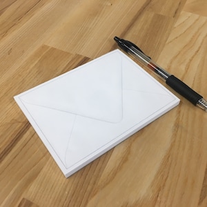 May include: A white envelope with a gray border and a black and clear pen with a black grip rest on a light brown wooden surface. The pen is positioned diagonally across the top right corner of the envelope.