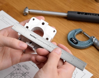 May include: A silver metal part is being measured with a ruler. The ruler is marked in inches and has the text "tarkka engineering and design" printed on it.