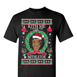 Pode incluir: Camiseta preta com um design de Natal com uma imagem de desenho animado do boxeador Mike Tyson usando um chap&eacute;u de Papai Noel e o texto "Kith Me Under The Mithletoe".