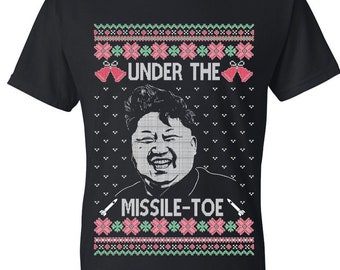 May include: Black t-shirt with a humorous holiday design. The shirt features a portrait and the text "UNDER THE MISSILE-TOE" in a festive, ugly sweater style. The design includes red, green, and white accents, and missile graphics.