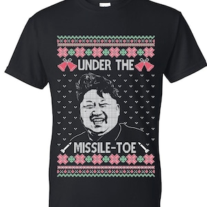 May include: Black t-shirt with a humorous holiday design. The shirt features a portrait and the text "UNDER THE MISSILE-TOE" in a festive, ugly sweater style. The design includes red, green, and white accents, and missile graphics.