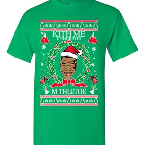 Pode incluir: Camiseta verde com um design de Natal com uma imagem de desenho animado do boxeador Mike Tyson usando um chap&eacute;u de Papai Noel e o texto "Kith Me Under The Mithletoe".