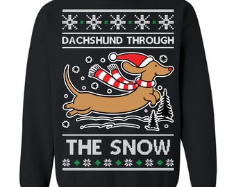 May include: Black sweatshirt with a dachshund wearing a Santa hat and scarf, jumping through the snow. The text "DACHSHUND THROUGH THE SNOW" is printed above and below the dachshund illustration. Snowflake and Christmas tree designs are also included.