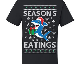 May include: Black t-shirt with a festive design featuring a blue shark wearing a Santa hat and holding a gift. The text "SEASON'S EATINGS" is displayed in white, with snowflake and decorative border elements.