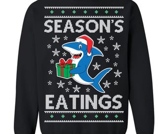May include: Black sweatshirt with a graphic of a blue shark wearing a Santa hat and holding a green gift box. The text "SEASON'S EATINGS" is displayed in white, with snowflake and decorative patterns.