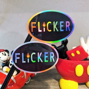 Könnte beinhalten: Zwei ovale, schimmernde Aufkleber mit dem Wort "FLICKER" in weißen Buchstaben auf schwarzem Hintergrund. Das Wort "FLICKER" ist mit einer Flammengrafik in der Mitte des Wortes stilisiert.