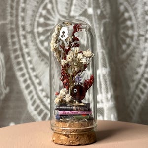 May include: A glass cloche with a cork base, containing dried flowers, miniature books, and a decorative object. The books have titles like "All Rhodes Lead Her" and "By a Thread". The decorative object is a dark red sphere with a purple gem on top.