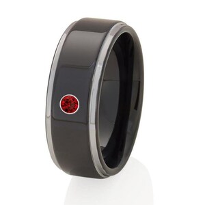 May include: A black ring with a silver trim and a single, round, red gemstone. The ring has a smooth, polished surface and a modern design. The gemstone is set in a circular bezel.