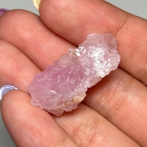 May include: A raw, pale pink crystal cluster with a rough, textured surface. The crystal is held in a hand, showcasing its natural form and colour variations. The crystal appears to be a mineral specimen.