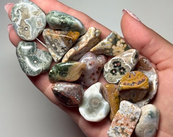 May include: A selection of polished gemstones in various shapes and sizes, held in a hand. The stones display a range of colours, including green, brown, white, and yellow, with unique patterns and textures. The gemstones are smooth and appear to be tumbled.