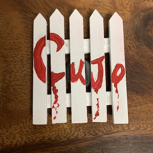 May include: A white picket fence with the word "CUTIE" painted in red. The fence is made of wood and has a distressed look. The background is a wooden surface.