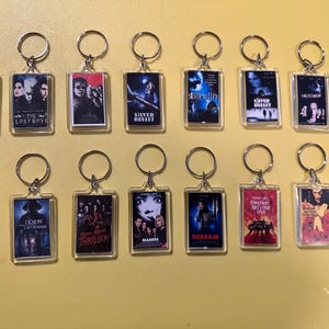 May include: A collection of rectangular keychains featuring movie posters. The keychains have silver-colored rings and clear plastic cases. The posters are for various horror films, including "The Lost Boys" and "Scream."
