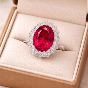 Puede incluir: Un anillo de plata con una gran gema roja oscura de corte ovalado, rodeada por un halo de piedras claras más pequeñas. El anillo se presenta en un joyero marrón con interior beige, creando una sensación lujosa.