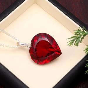 May include: A heart-shaped, faceted red pendant necklace with a silver chain. The pendant is displayed in a black box with a cream-colored interior, accented by a sprig of green foliage.
