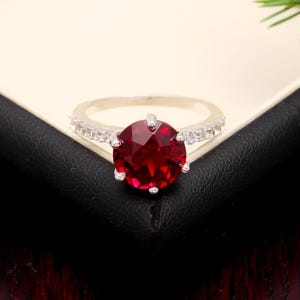 Op de afbeelding: Een zilveren ring met een grote, facetgeslepen, dieprode edelsteen als middelpunt. De band is versierd met kleinere, heldere edelstenen. De ring ligt op een zwart oppervlak, met een witte en beige achtergrond.