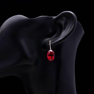 May include: A single red oval-cut gemstone earring on a silver hook. The earring is displayed on a black mannequin head. The gemstone has a faceted surface, reflecting light. The earring is a classic jewelry piece.