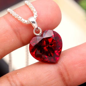 May include: A silver necklace featuring a heart-shaped, faceted, deep red pendant. The pendant is a vibrant gemstone. The chain is a delicate, square-link style, adding elegance to the jewellery.