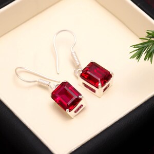 Può includere: Un paio di orecchini pendenti in argento con pietre preziose rettangolari rosso intenso. Gli orecchini sono incastonati in una montatura d'argento e hanno un semplice design a gancio. Gli orecchini sono esposti in un portagioie bianco.