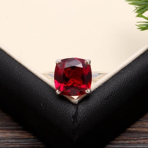 Può includere: Un anello d'argento con una grande gemma rosso scuro a taglio quadrato. La gemma è incastonata con quattro griffe. L'anello è presentato in un cofanetto nero. Un gioiello suggestivo.