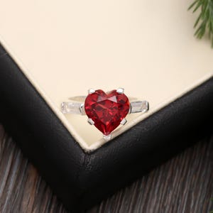 May include: A silver ring with a large, red heart-shaped gemstone. The ring has smaller clear gemstones on either side of the heart.