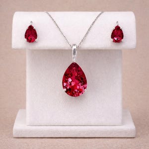 Puede incluir: Un conjunto de joyas con un collar con colgante y pendientes a juego. El colgante y los pendientes muestran grandes piedras preciosas de color rojo rubí en forma de lágrima. El collar tiene una delicada cadena de plata.