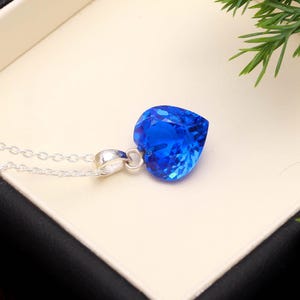 May include: A vibrant blue, heart-shaped pendant necklace with a faceted design. The pendant hangs from a delicate silver chain. The necklace is presented on a cream surface, with a dark border and green foliage in the background.