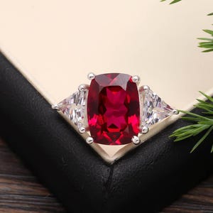 Könnte beinhalten: Ein silberner Ring mit einem großen, rechteckigen, tiefroten Edelstein im Zentrum, flankiert von zwei kleineren, dreieckigen, klaren Edelsteinen. Der Ring ist vor einem neutralen Hintergrund mit etwas Grün platziert.