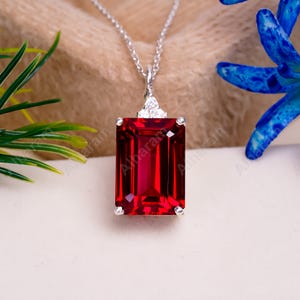 Könnte beinhalten: Eine Halskette mit einem roten Edelstein-Anhänger und einer silbernen Kette. Der Edelstein ist im Smaragdschliff geschliffen und von kleineren Diamanten umgeben.