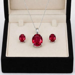 Può includere: Un set di gioielli con una collana d'argento con un grande pendente ovale in pietra rossa e orecchini a bottone ovali in pietra rossa abbinati. Il set è esposto in un cofanetto nero con interno color crema.