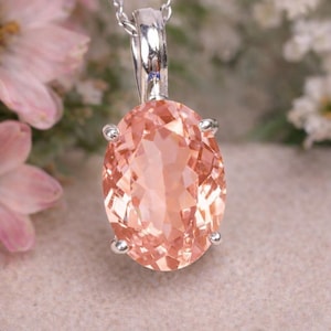 Op de afbeelding: Een zilveren hangerketting met een grote, ovaal geslepen perzik-kleurige edelsteen. De edelsteen is gezet in een zilveren bezel met vier klauwen. De ketting is ook zilver. De achtergrond is vervaagd met roze en witte bloemen.