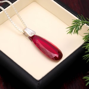Puede incluir: Un collar de plata con un colgante facetado en forma de lágrima de color rojo intenso. El colgante se muestra en una caja color crema con un borde negro, con follaje verde en el fondo.