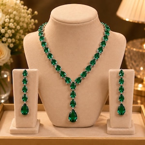 May include: A jewelry set consisting of a necklace and a pair of drop earrings. The necklace features a series of pear-shaped, deep green stones set in silver-toned metal, forming a V-shape with a central drop. Each earring has four matching stones.