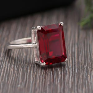 Puede incluir: Un anillo de plata con una gran gema rectangular de color rojo intenso. La gema es el punto focal, engastada en una banda simple y elegante. El anillo se exhibe sobre una superficie de madera oscura.