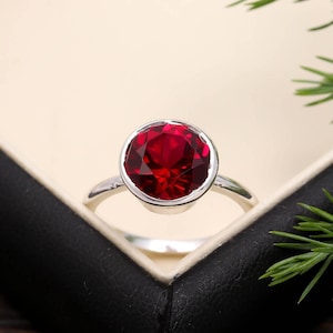 May include: A silver ring featuring a large, round, faceted, deep red gemstone. The ring has a bezel setting and a thin, polished band. It's presented in a black box with a cream interior, with green sprigs in the background.