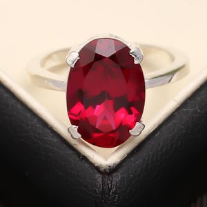 Puede incluir: Un anillo de plata con una gran gema roja oscura de corte ovalado. La gema es el punto focal, engastada en un engaste clásico de cuatro puntas. La banda del anillo es de plata pulida. El anillo se muestra sobre una superficie negra.