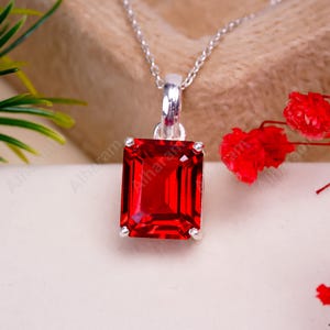 May include: A silver chain necklace with a red gemstone pendant. The gemstone is a square cut with beveled edges.