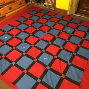 May include: A red and blue checkered quilt with black squares. The quilt is made of fabric squares sewn together. The quilt is on a carpeted floor.