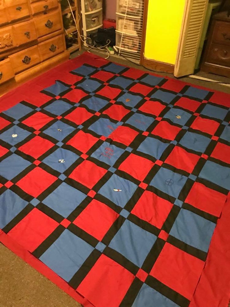 May include: A red and blue checkered quilt with black squares. The quilt is made of fabric squares sewn together. The quilt is on a carpeted floor.