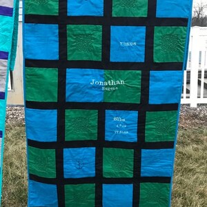 May include: A blue and green quilt with black squares. The quilt has embroidered details, including a baseball, a date, a name, and a weight and measurements. The quilt is hanging on a clothesline.