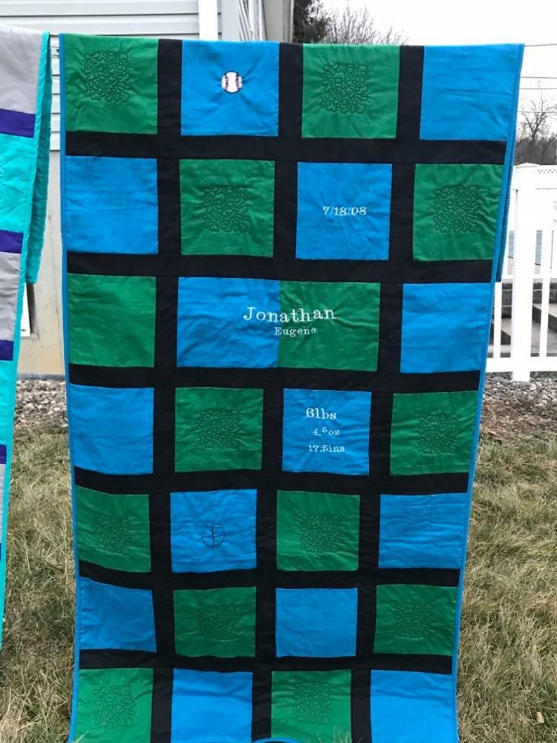 May include: A blue and green quilt with black squares. The quilt has embroidered details, including a baseball, a date, a name, and a weight and measurements. The quilt is hanging on a clothesline.