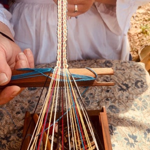 May include: A close-up of a wooden loom with colorful threads being woven into a pattern. The threads are arranged in a radial pattern, with the center of the pattern being the top of the loom. The loom is made of wood and has a simple design.