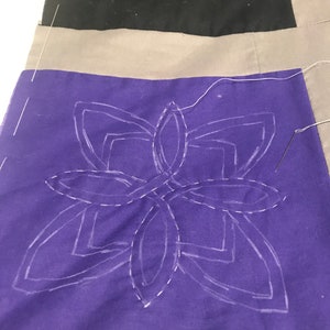 May include: A purple fabric square with a white outline of a geometric design. The design is a series of overlapping circles and squares. The fabric is being stitched with white thread.