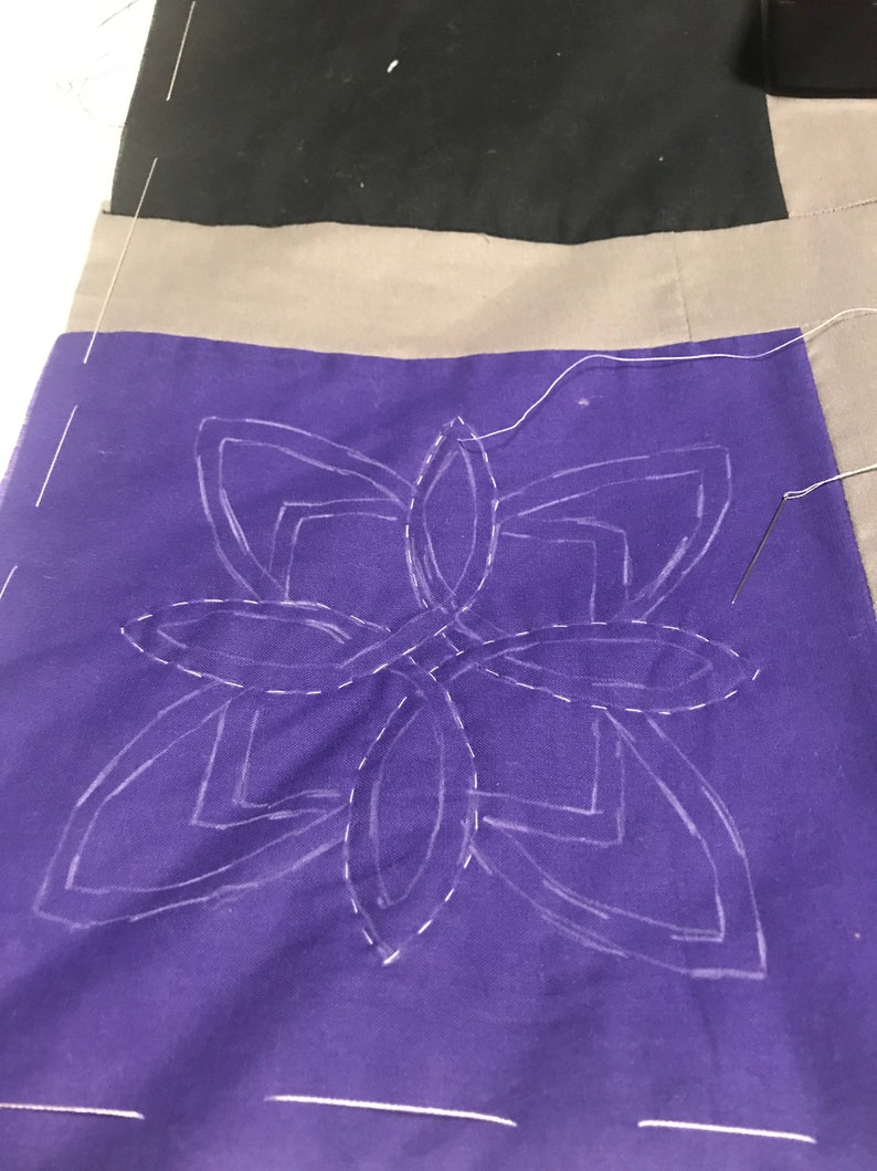 May include: A purple fabric square with a white outline of a geometric design. The design is a series of overlapping circles and squares. The fabric is being stitched with white thread.
